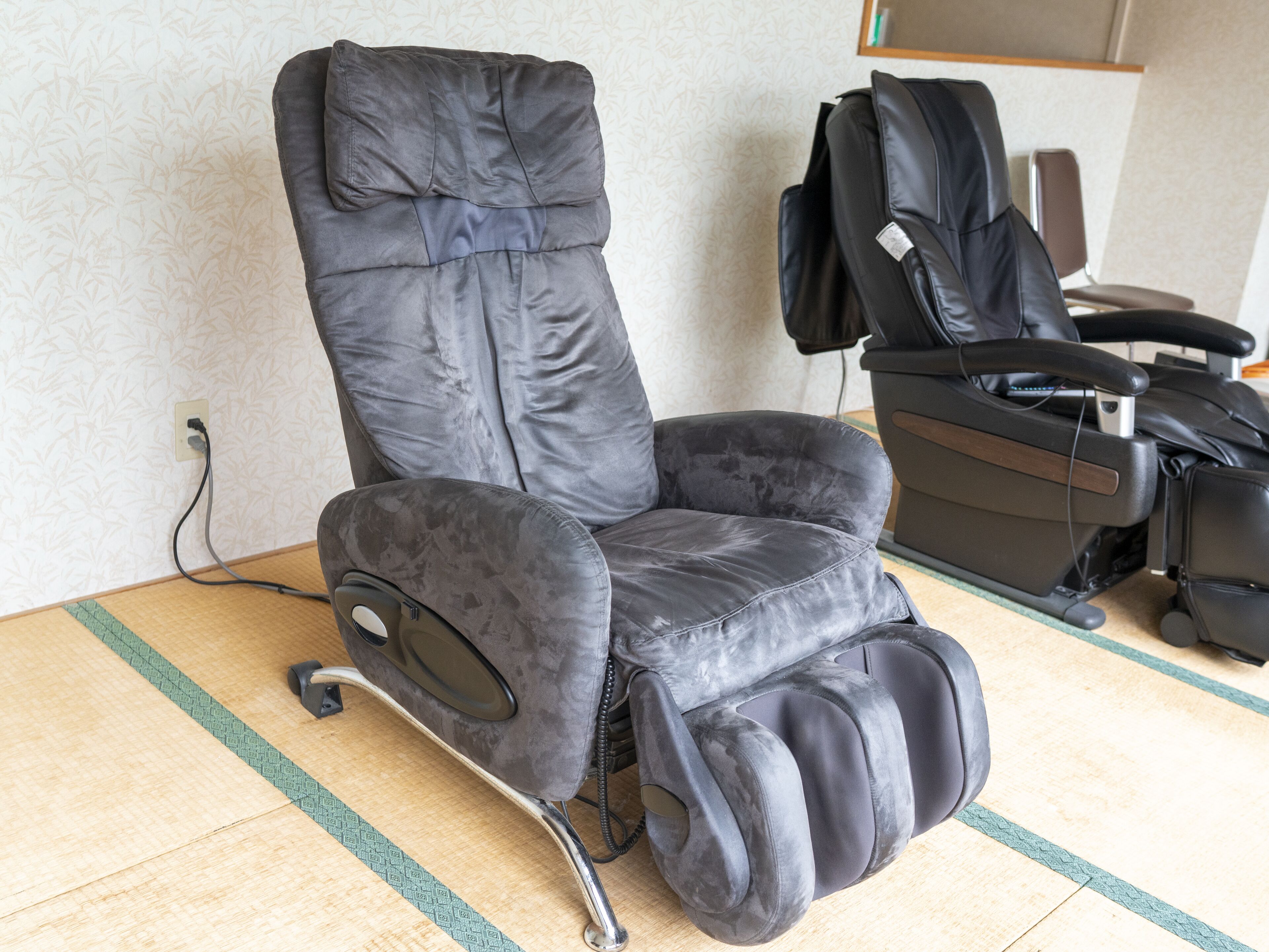 Een massagestoel met beensteun en bedieningspaneel, geplaatst op een houten vloer met een kamerkleed met patroon.