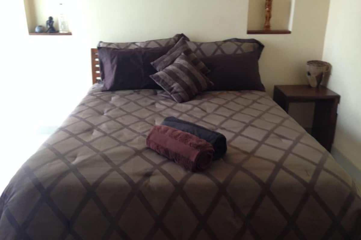 A neatly made bed with a patterned bedspread and several pillows, a small bedside table with a lamp, and a shelf with decorative items.