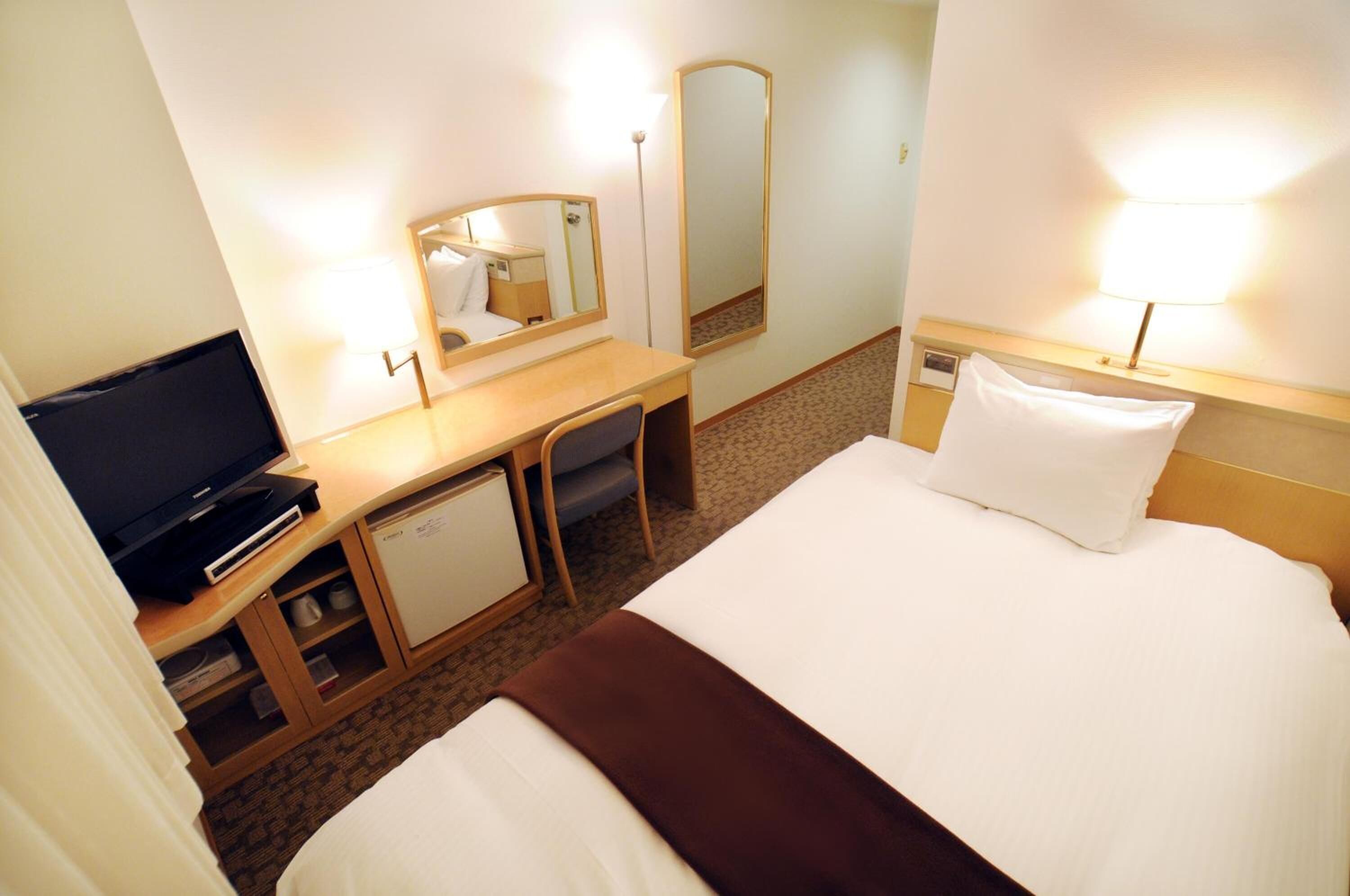 Habitación de hotel con cama, escritorio, televisor, espejo y lámpara.