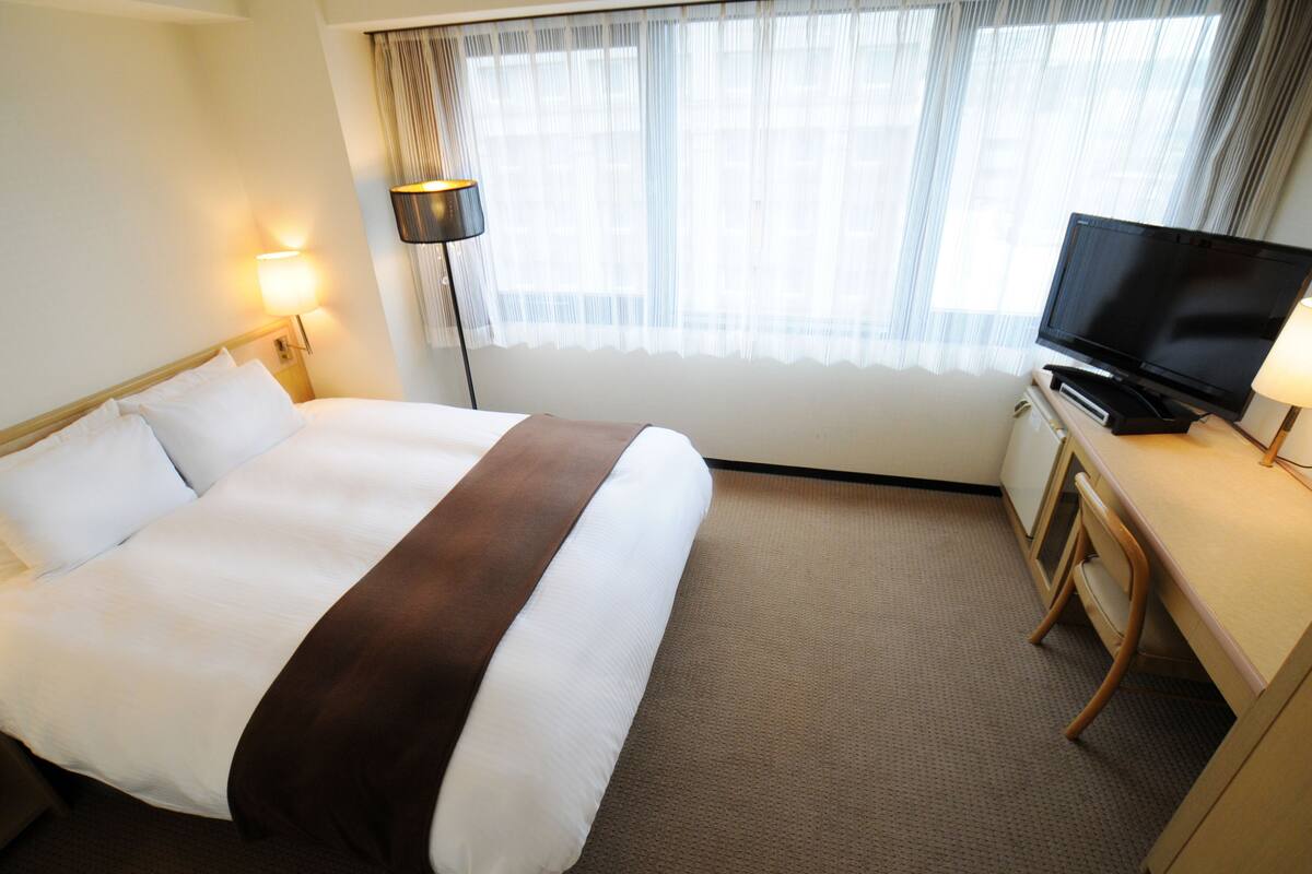Une chambre d’hôtel avec un lit, un bureau avec une télévision, une lampe et une fenêtre avec des rideaux.