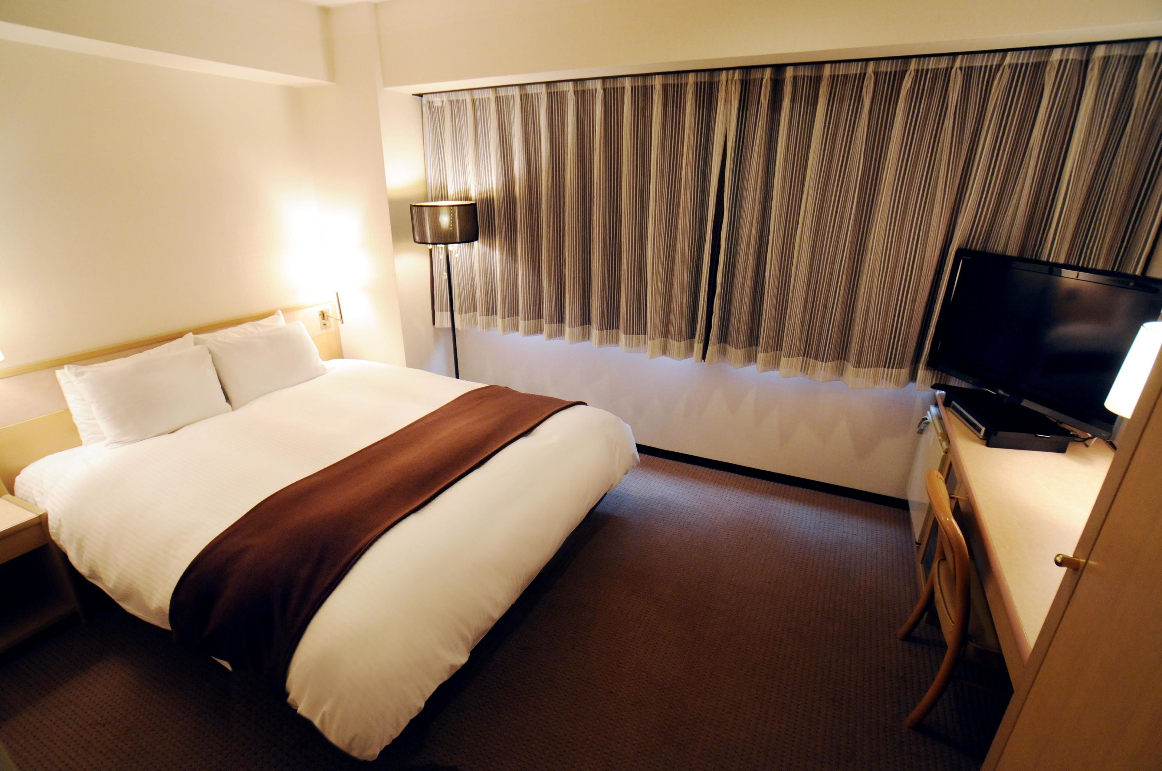 A hotel room with a large bed, a flat-screen TV, a desk with a chair, and a window with curtains.