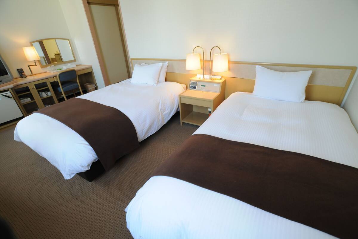 Une chambre d’hôtel avec deux lits simples, un bureau avec une chaise, une table de chevet avec des lampes et un miroir.