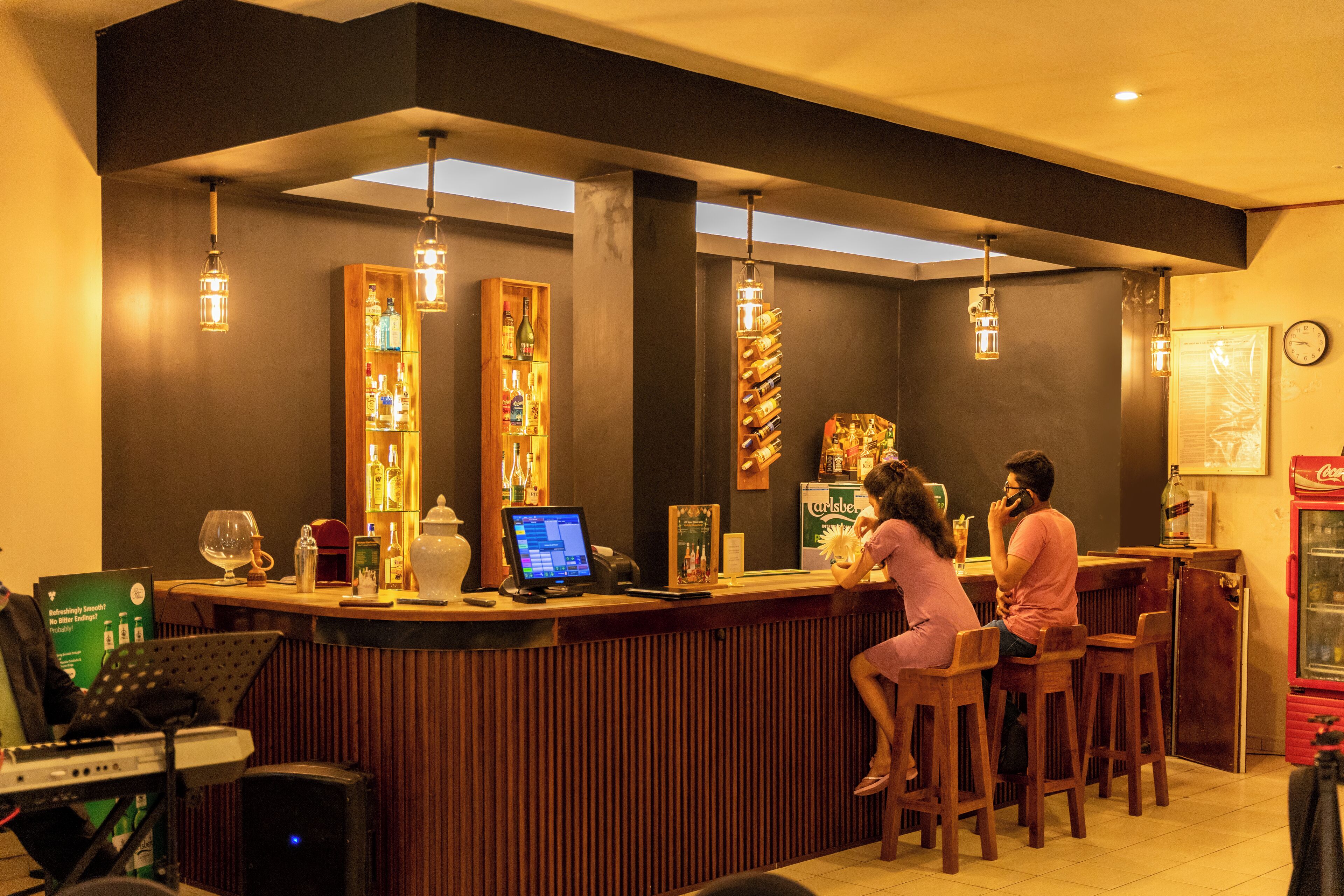 A bar area with wooden stools, a bar counter, and a computer monitor. There are shelves with bottles and a clock on the wall.