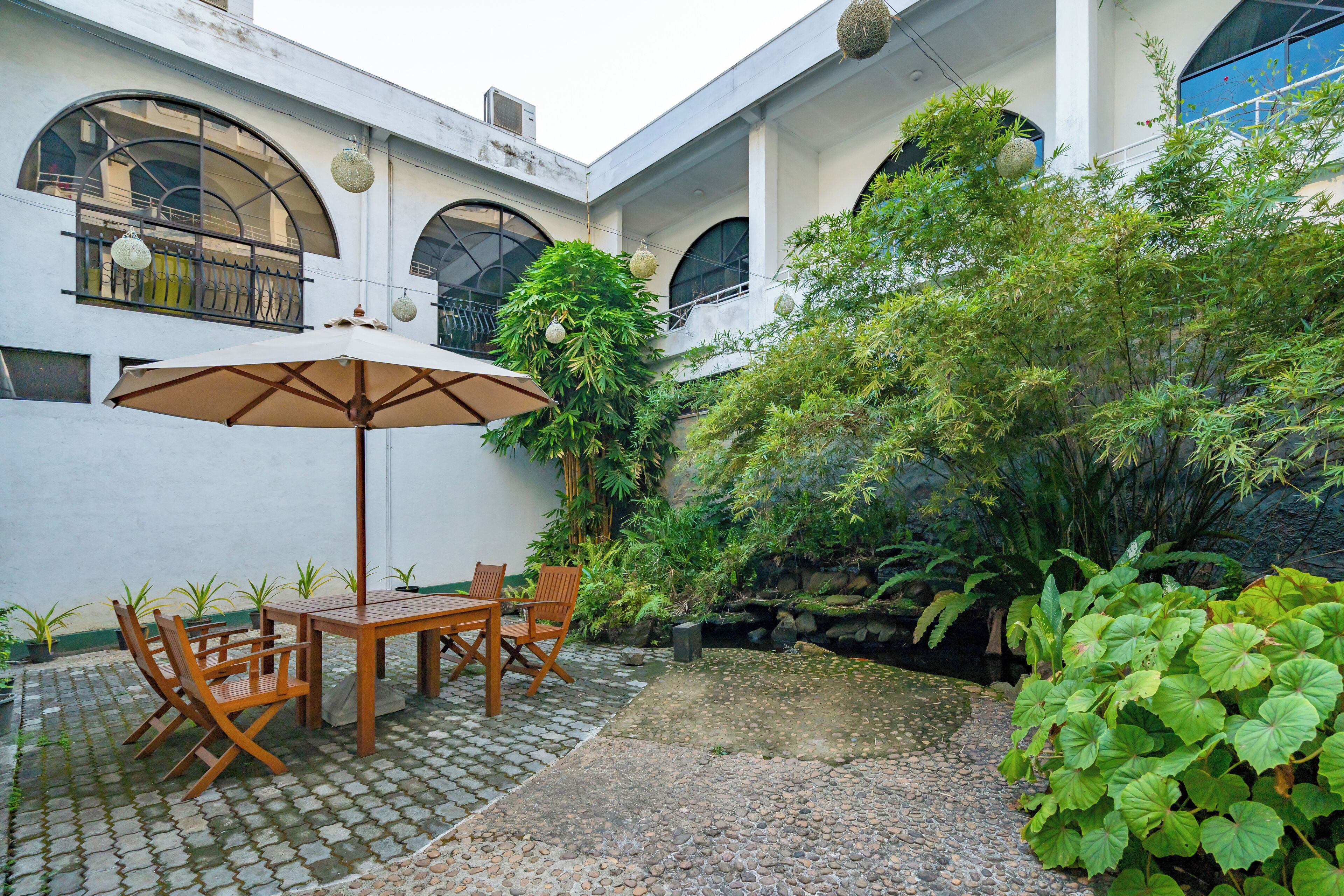 A courtyard with a wooden table and chairs, surrounded by lush greenery and a stone pathway.