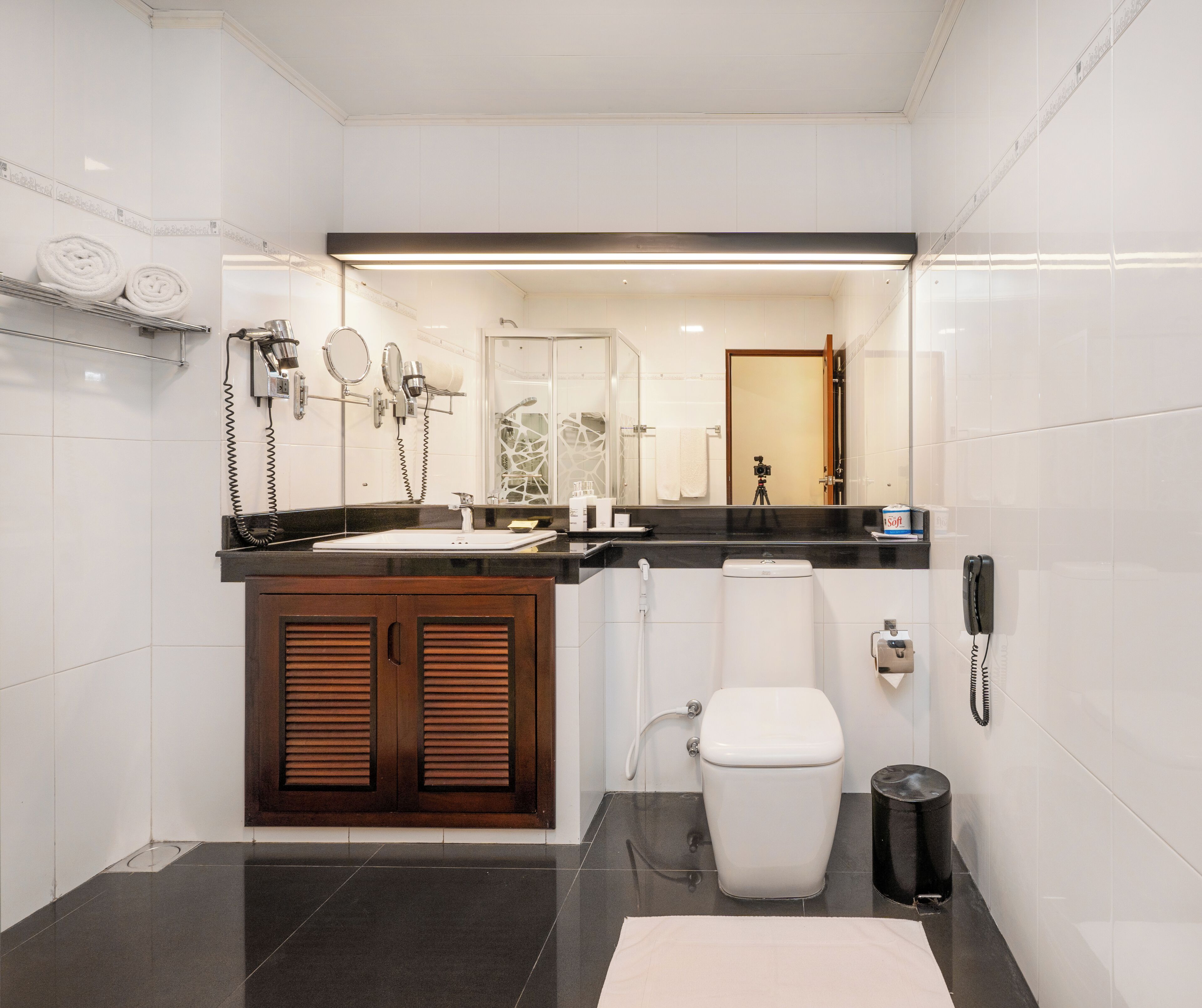 A modern bathroom with a black countertop, white tiles, a wooden cabinet, a toilet, and a trash can.