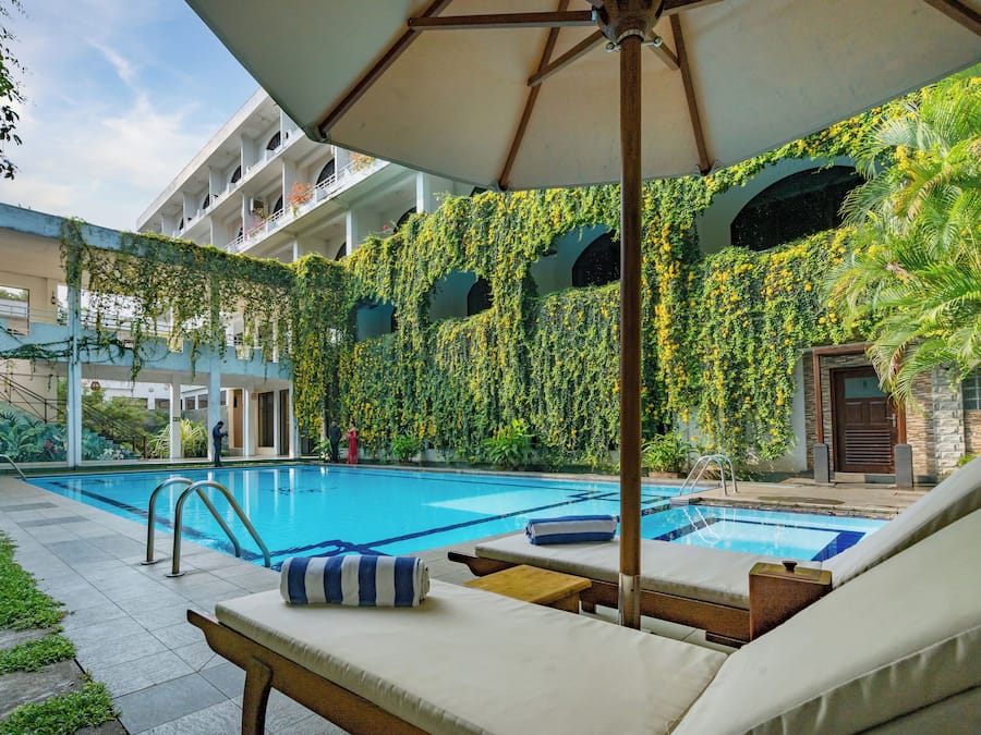 A swimming pool with lounge chairs and a covered area, surrounded by greenery and a building with multiple balconies.