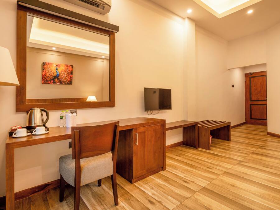 A hotel room with a wooden desk, a chair, a television, a painting, and a wooden bench.