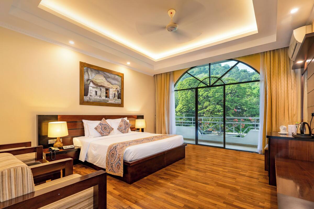 A hotel room with a large bed, two bedside tables with lamps, a wooden floor, a painting on the wall, and a large window with a view of greenery.