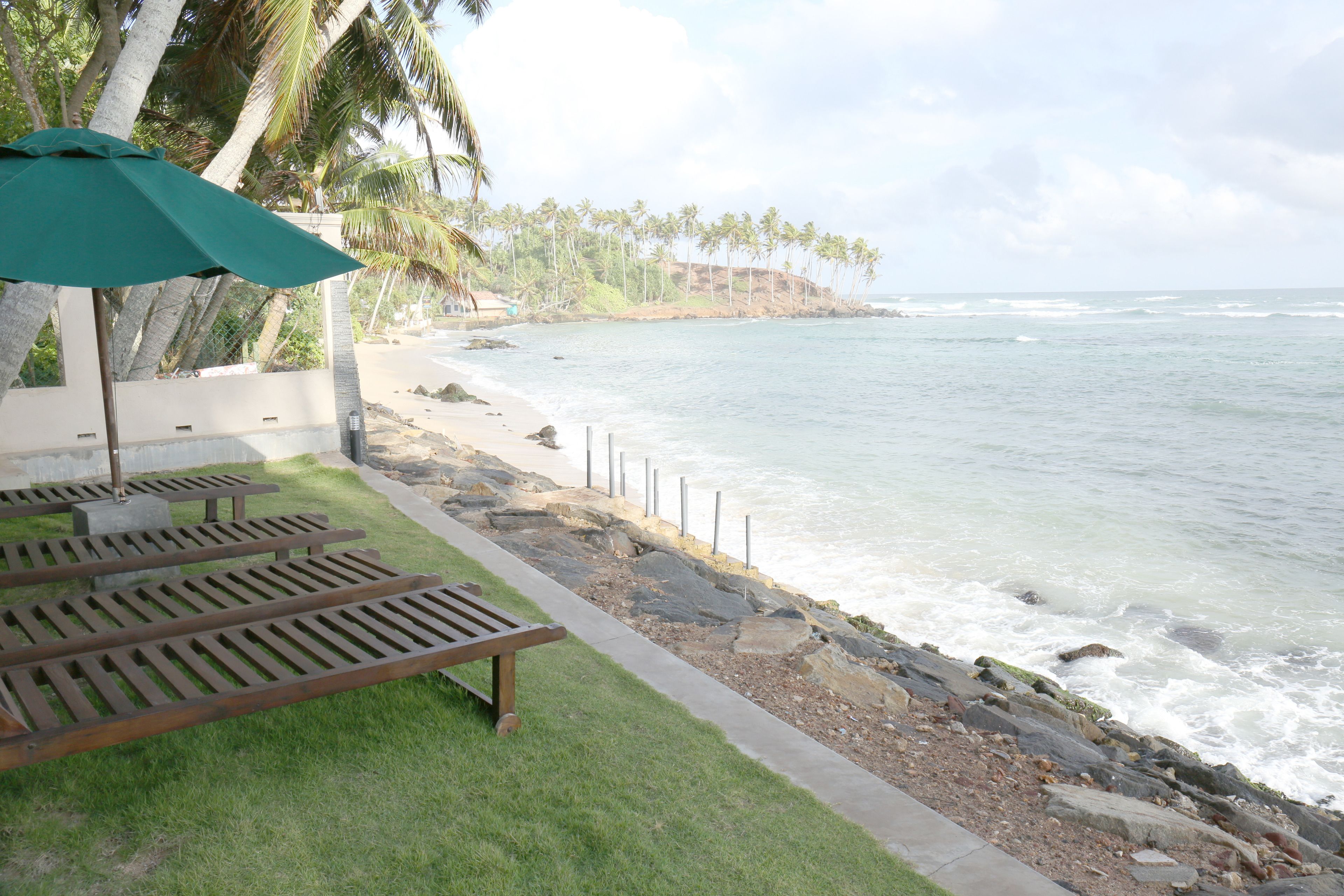 A beachside area with wooden loungers and a green umbrella, a concrete pathway, and a rocky shoreline with palm trees in the background.