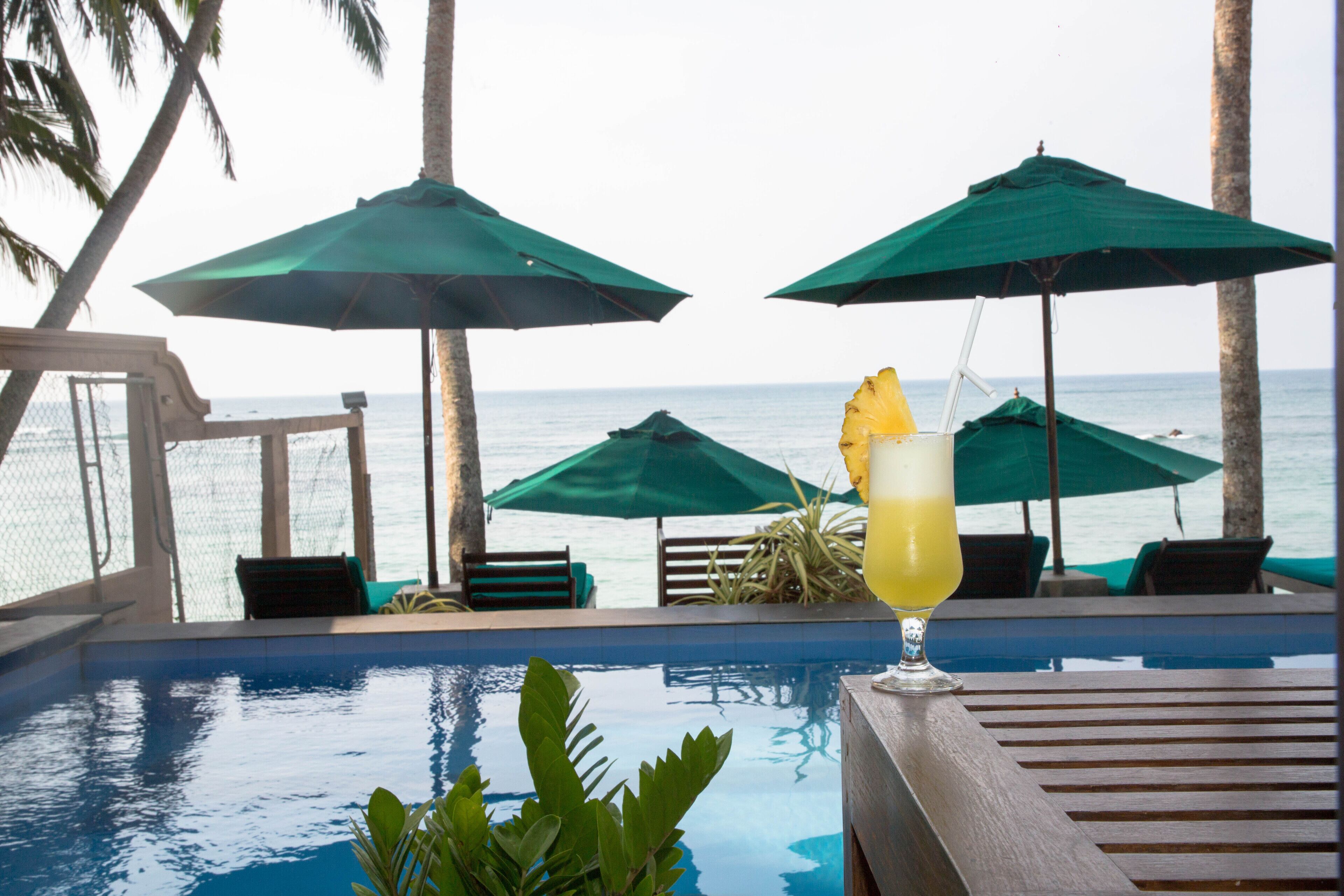 An indoor pool with a view of the ocean, surrounded by palm trees and green umbrellas.