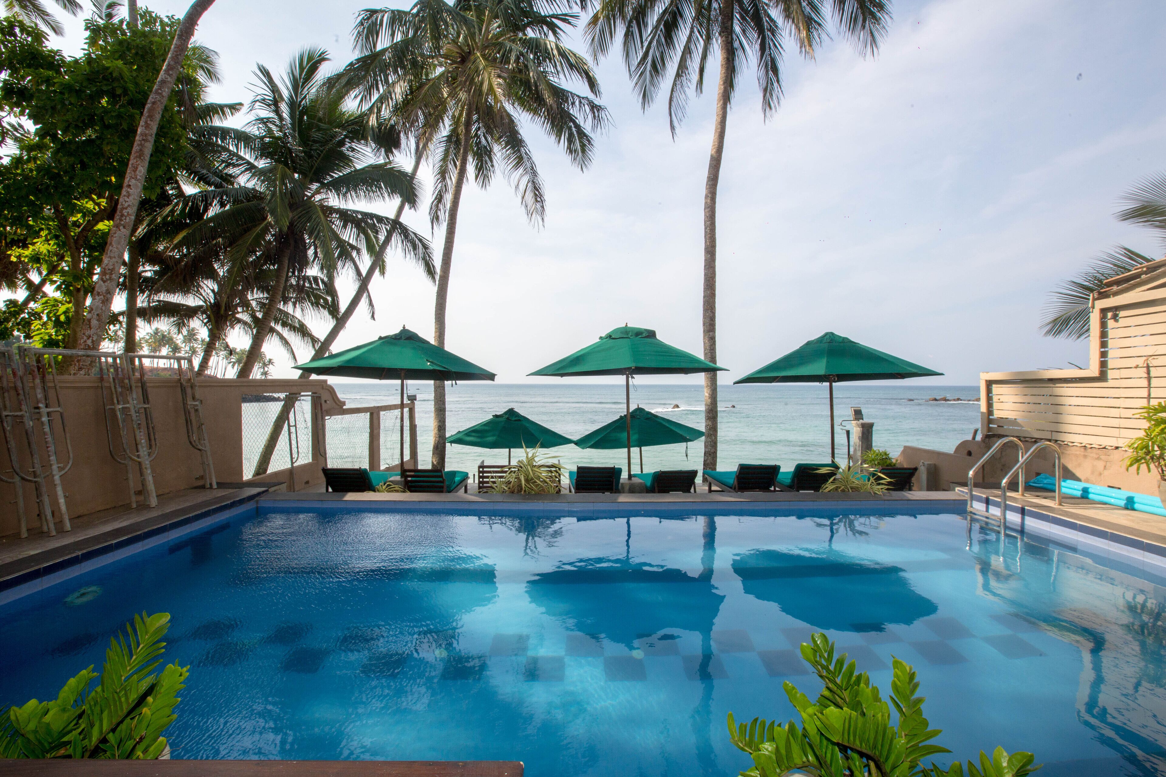 A swimming pool with lounge chairs and umbrellas, surrounded by palm trees and a view of the sea.