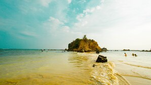 Uma praia com água cristalina, uma ilha rochosa e pessoas nadando.