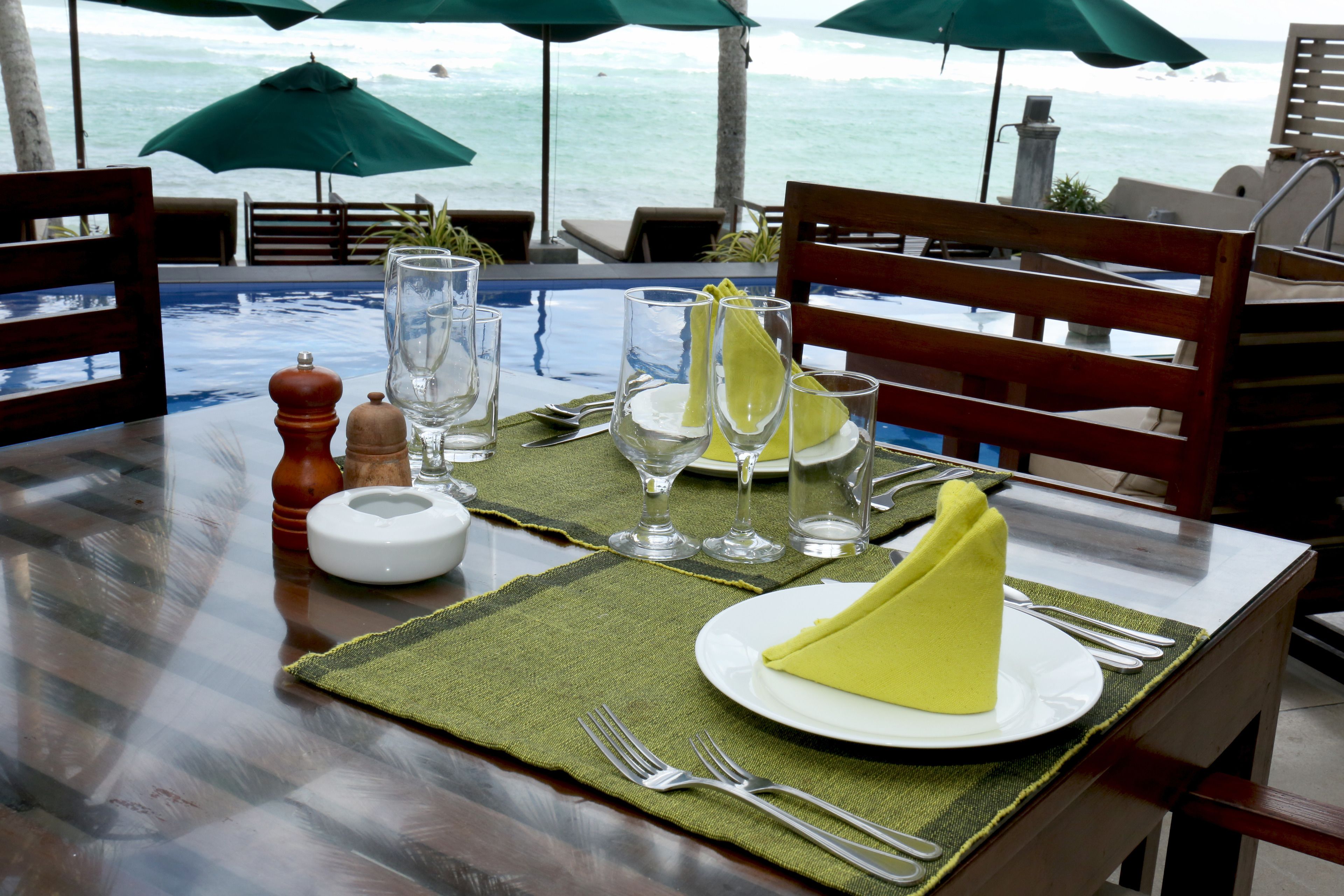 A table set for dining with a view of the ocean, equipped with green umbrellas, and a wooden chair.