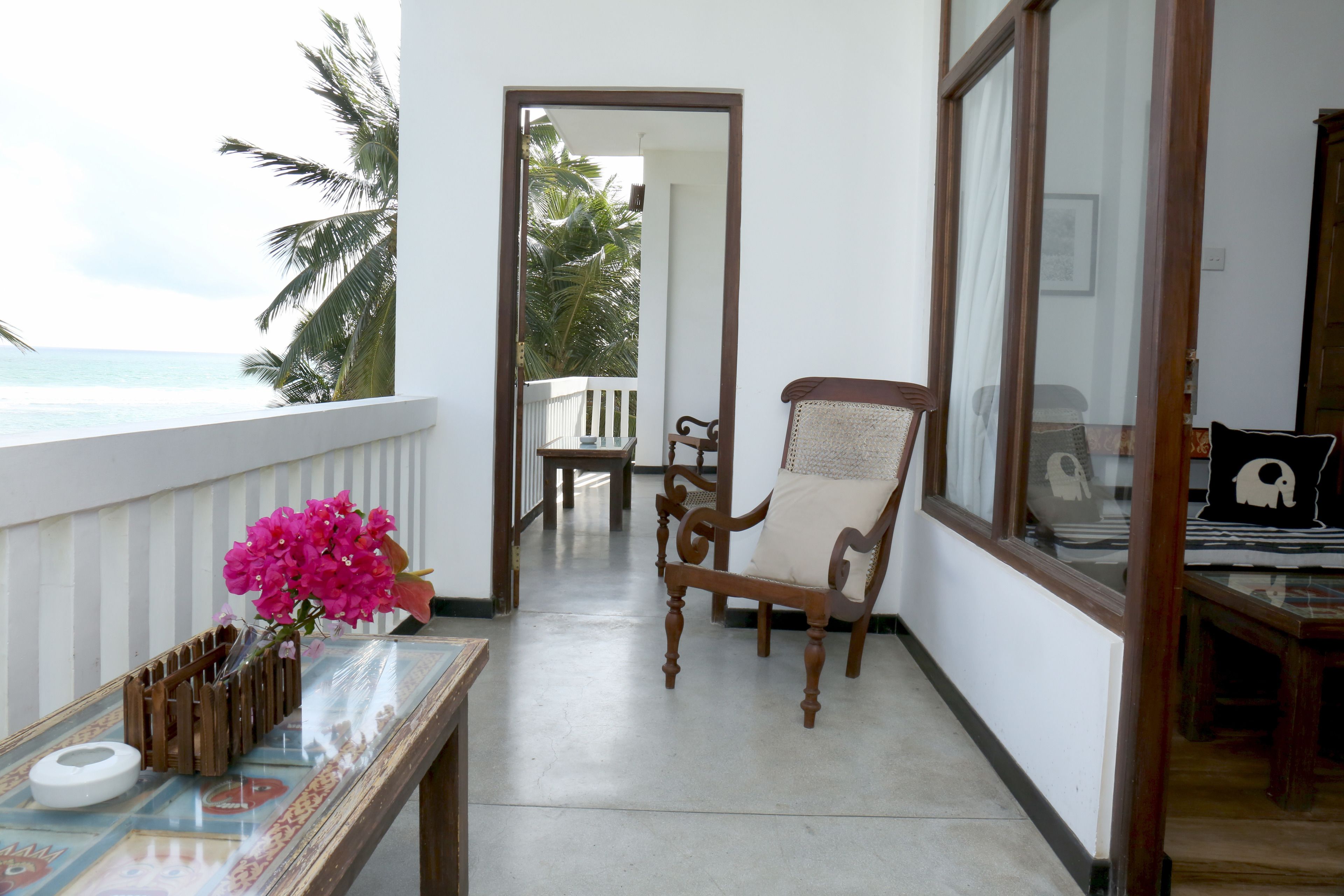 A balcony with a view of the ocean, a wooden chair, a table with a vase of flowers, and a glimpse of a room with a bed and a framed picture.