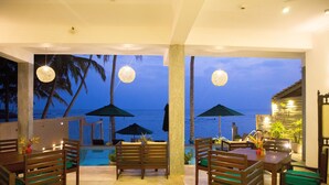 Área de refeições ao ar livre com mobiliário de madeira e guarda-sóis, com vista para uma piscina e palmeiras ao entardecer.