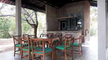 An outdoor covered seating area with wooden chairs and a table, a bar with shelves of bottles, and a tree in the background.
