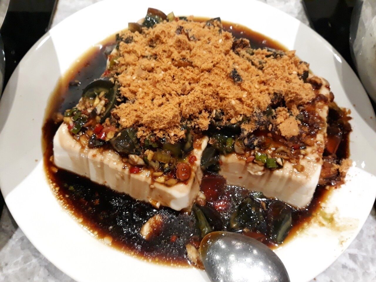 A dish with tofu, vegetables, and a dark sauce, served on a white plate with a spoon.