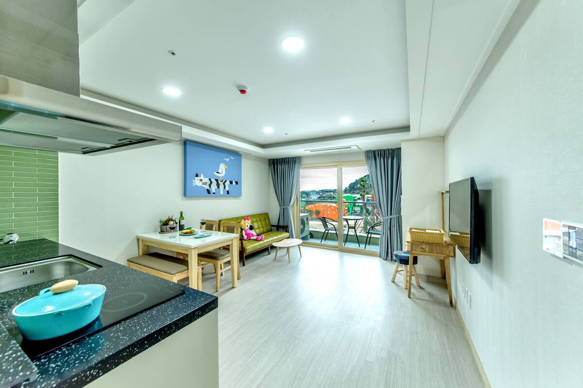 A modern kitchen with a blue bowl on the counter, a dining area with a table and chairs, a sofa, and a TV mounted on the wall.