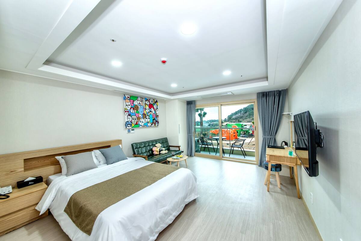 A modern hotel room with a large bed, a desk with a chair, a TV mounted on the wall, and a balcony with a view of the city.