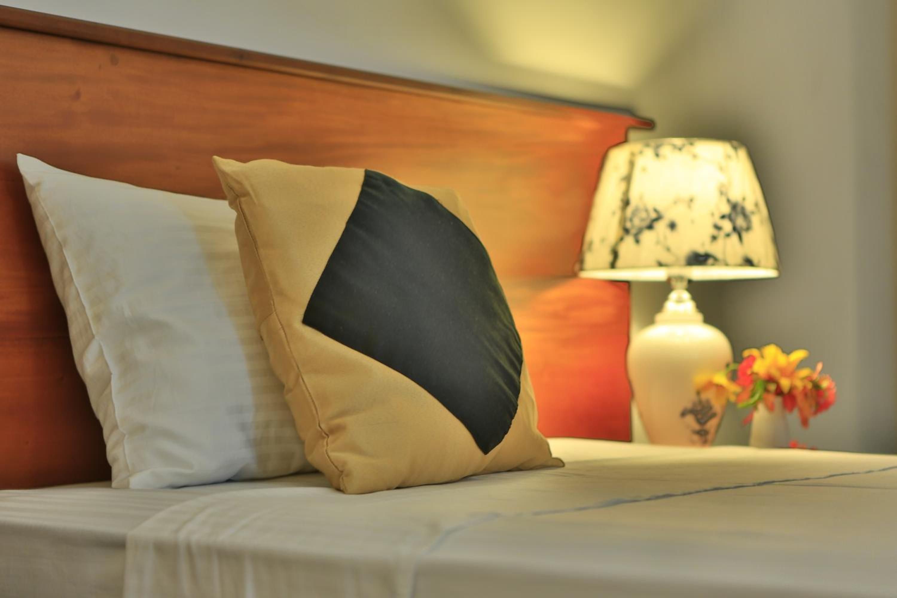 Uma cama com duas almofadas, uma mesa de cabeceira com abajur e um vaso com flores.
