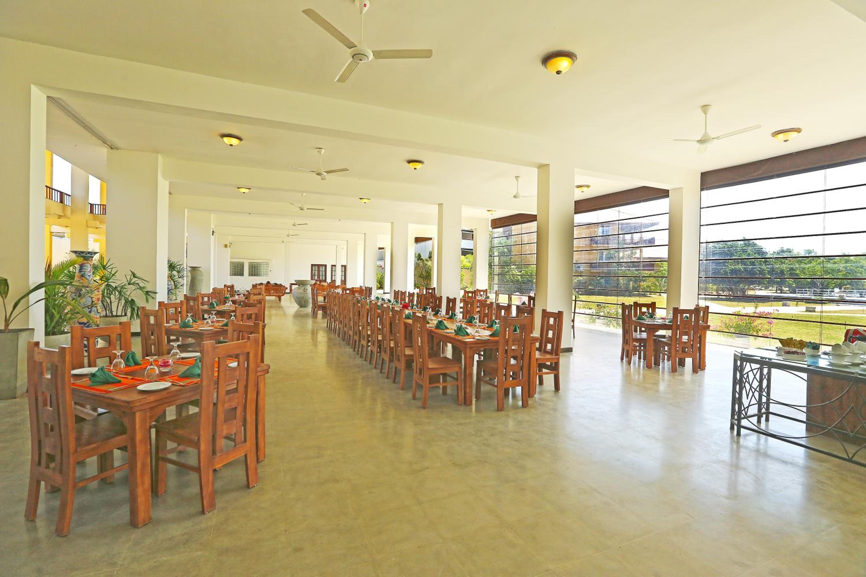 A spacious dining hall with multiple tables and chairs, large windows offering a view of the outdoors, and ceiling fans.