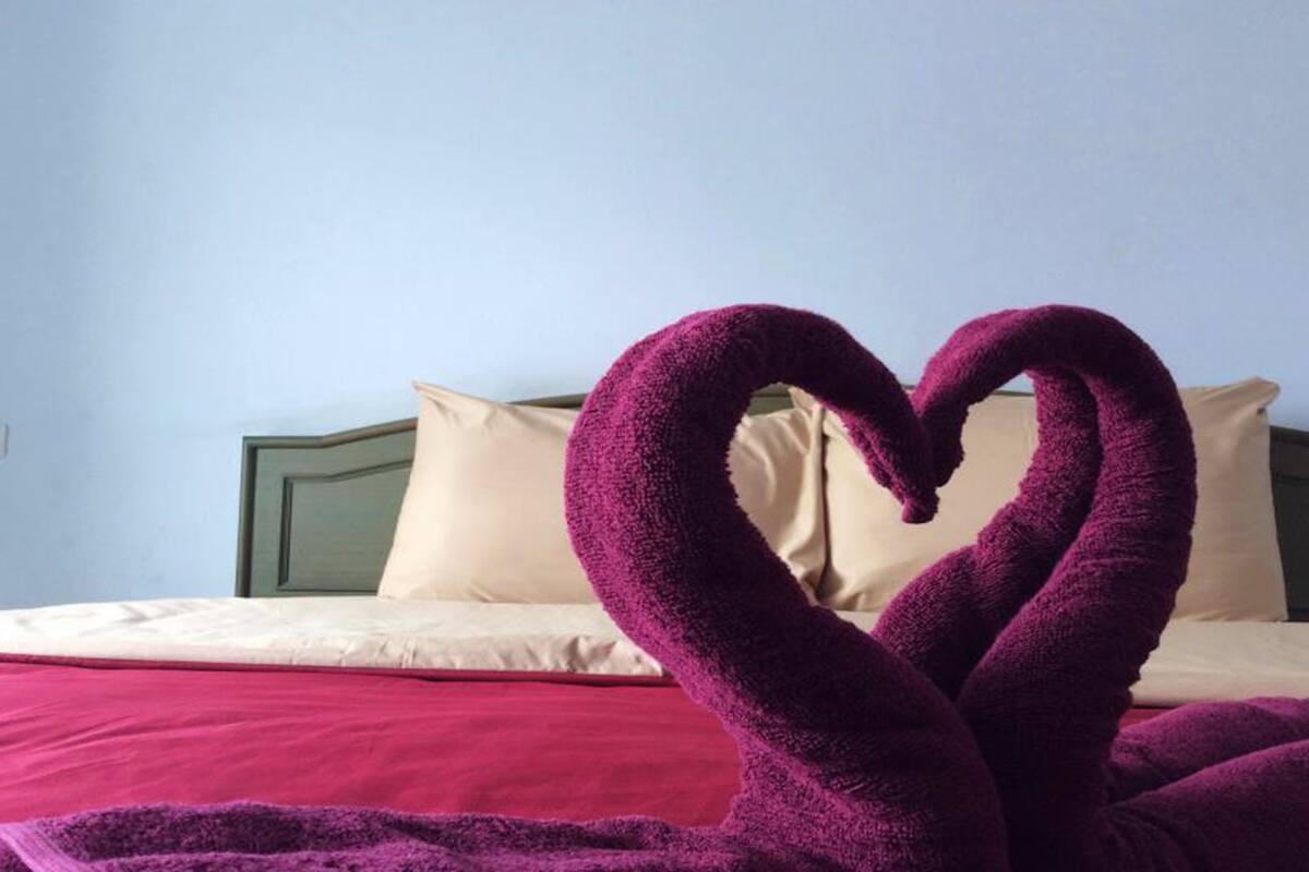Ein Bett mit roter Decke und Kissen, sowie zwei violette Handtücher, die in Herzform drapiert sind.