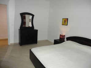 A bedroom with a bed, a dresser, a mirror, a bedside table with a lamp, and a painting on the wall. - Appartement LaVie (Tunis)