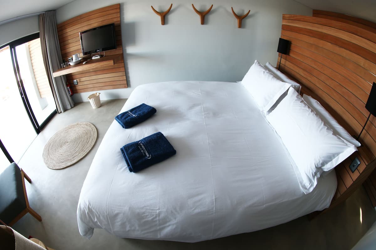 Ein modernes Hotelzimmer mit einem großen Bett, einem flachbildfernseher an der Wand, einem Holzkopfende und Blick ins Freie durch das Fenster.