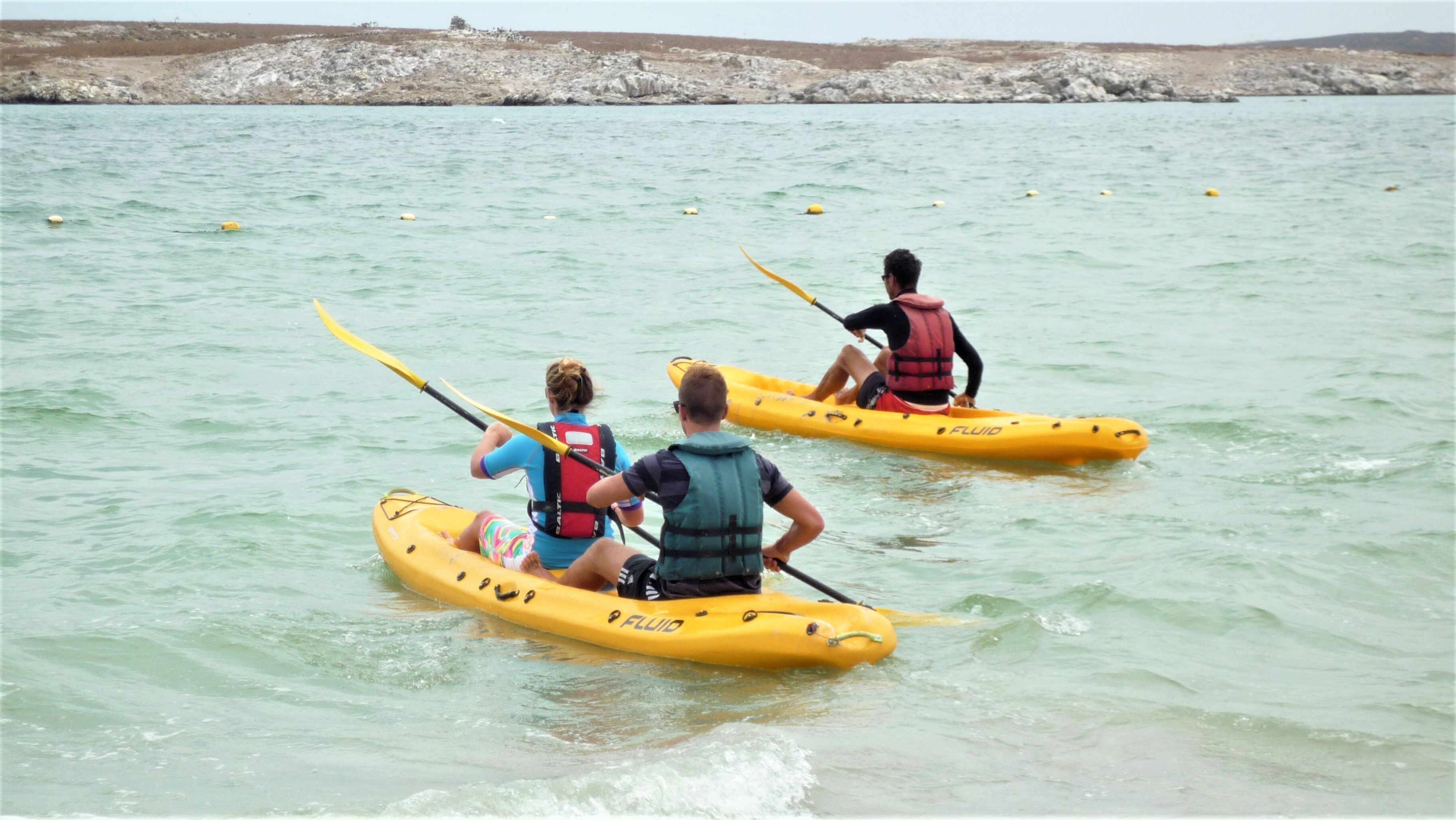 Tres personas haciendo kayak en aguas tranquilas con una costa rocosa al fondo.