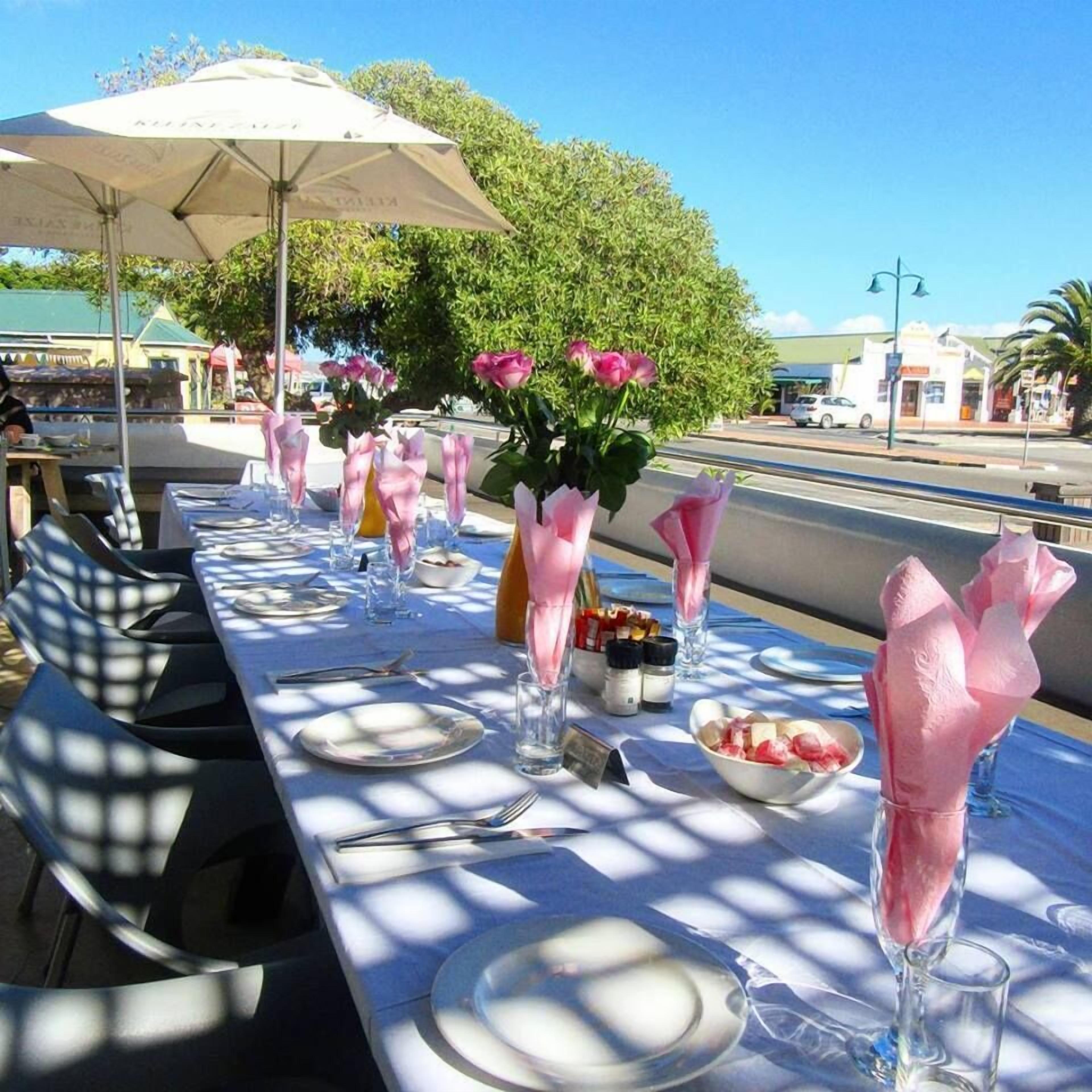 An outdoor dining setup with tables set for two, featuring pink napkins and clear glassware.