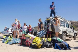 Un grupo de personas de pie cerca de un vehículo con tablas de surf y bolsas en el suelo.