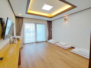 A hotel room with a wooden floor, a flat-screen TV on a wooden desk, a bed with white linens, a window with curtains, and a ceiling light. - Fellisia Hotel (Seogwipo)