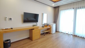A hotel room with a wooden desk, a flat-screen TV, a small refrigerator, a trash bin, and a large window with curtains. - Fellisia Hotel (Seogwipo)