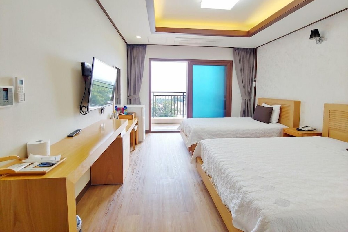 A hotel room with two beds, a desk with a computer, a flat-screen TV, and a balcony with a view.