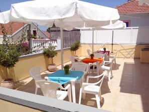 Un patio al aire libre con sillas y mesas de plástico blanco, algunas cubiertas por sombrillas blancas, y plantas en macetas.