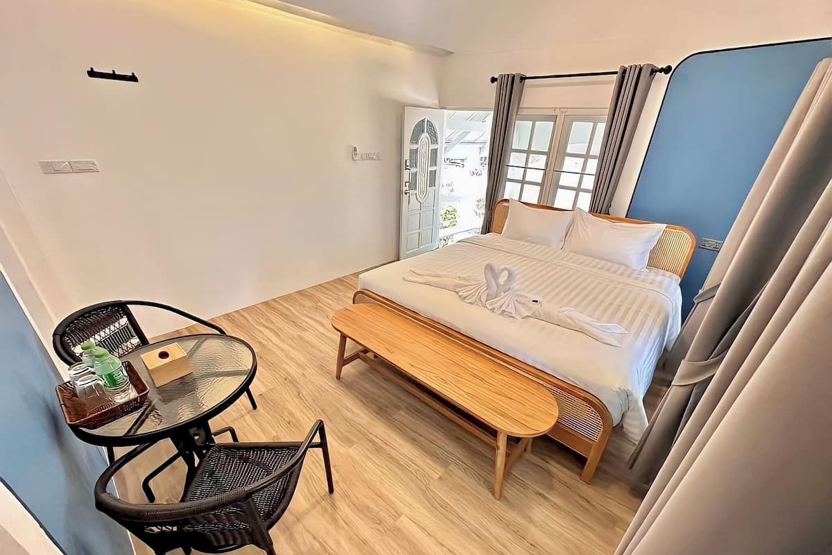 A compact hotel room with a bed, a small table with chairs, a bench, and a window with curtains.