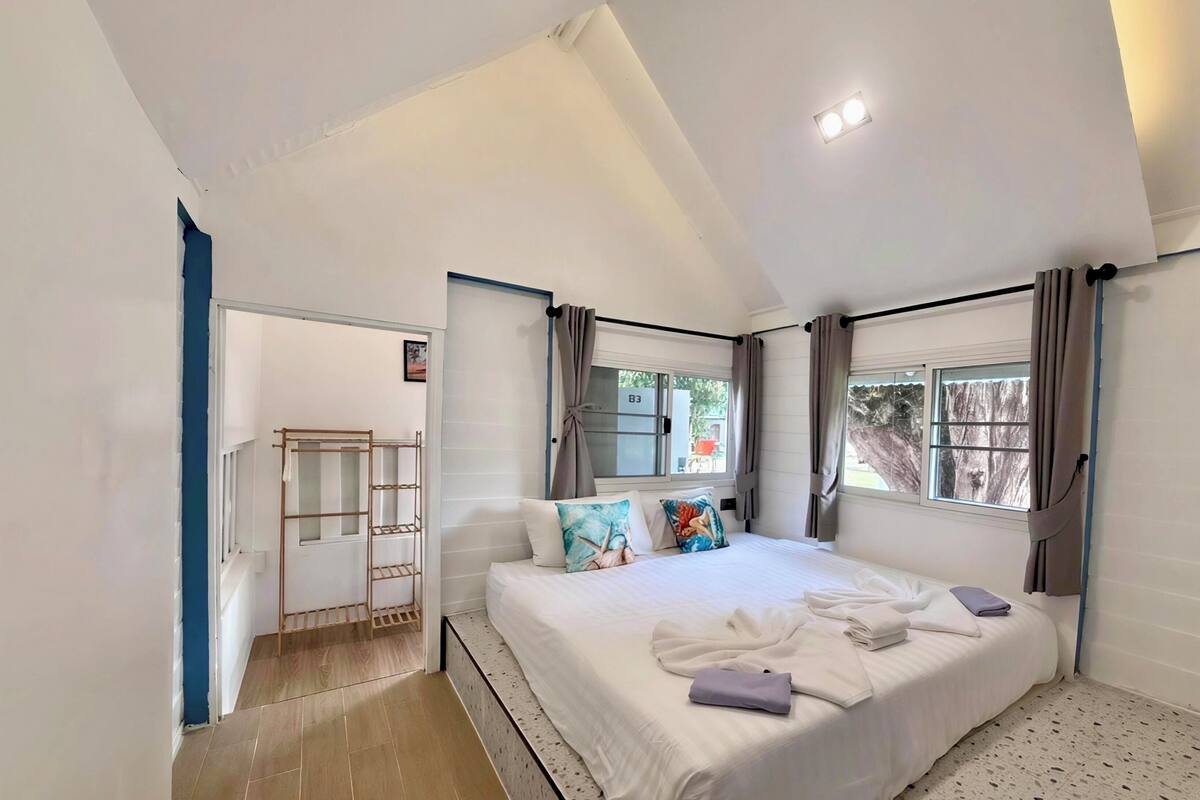 A bedroom with a large bed, a window with curtains, a wooden ladder, and a door leading to another room.