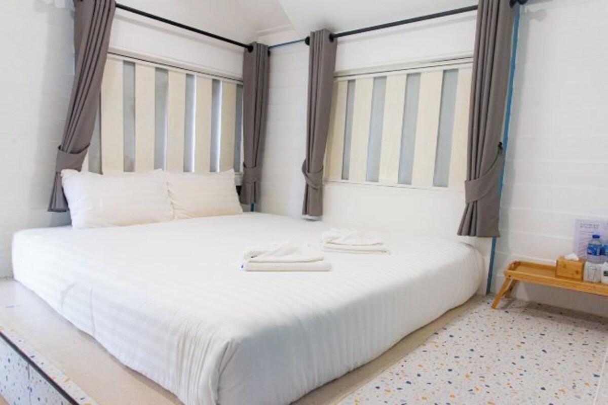 A neatly made bed with white linens, a wooden bedside table with a bottle and a tissue box, and a small wooden tray with toiletries.