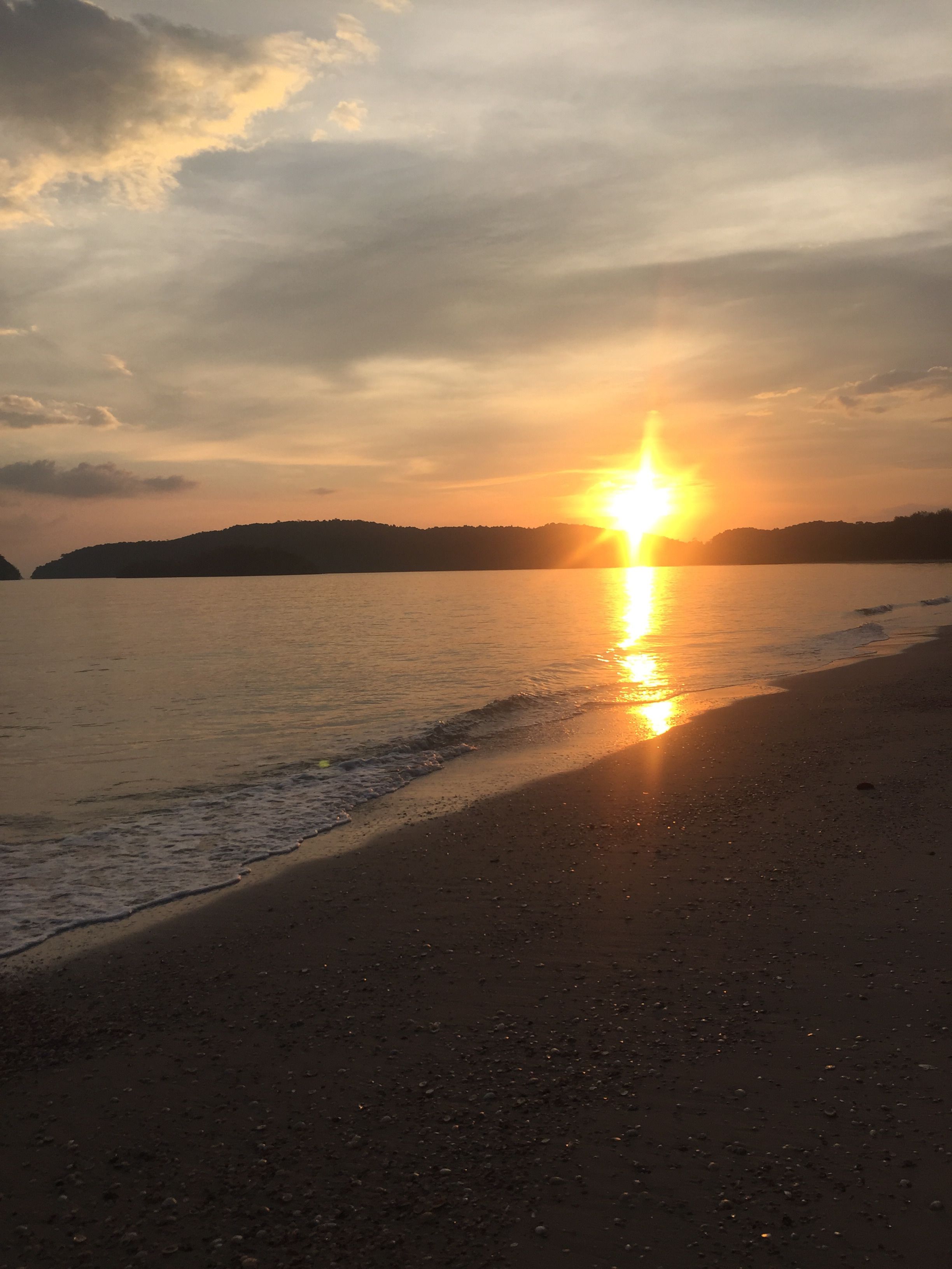 A beach with a sunset over water, a sandy shore, and distant hills.
