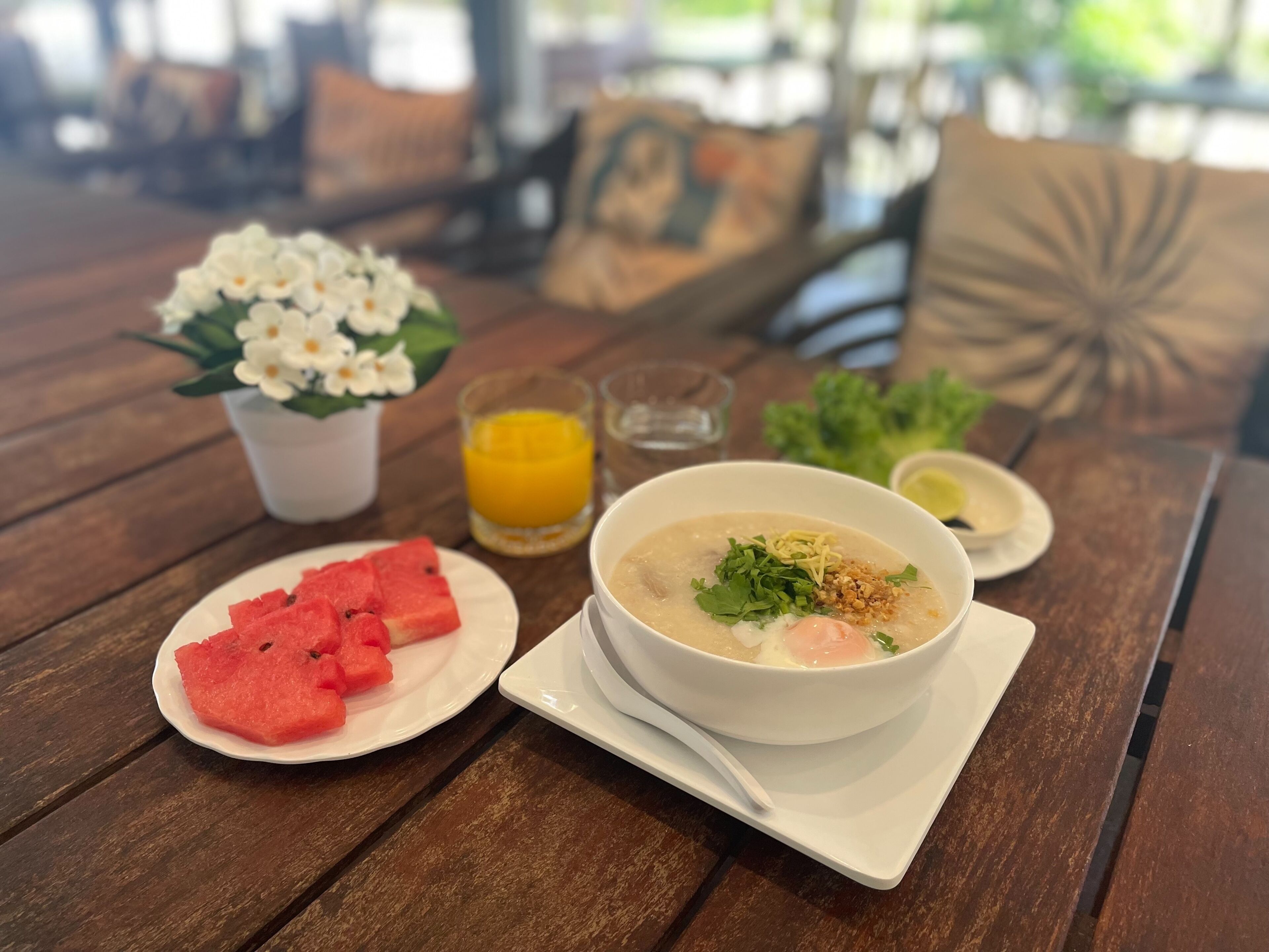 Une table avec un bol de soupe, des tranches de pastèque, un verre de jus d’orange et une petite assiette de légumes verts.