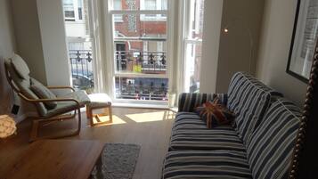 A living room with a striped sofa, a wooden chair, a small table, and a large window with a view of buildings and a clock.