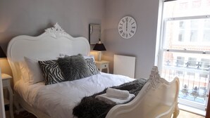 A bedroom with a white ornate bed, a round wall clock, a window with a view of buildings, and a radiator. - Brighton Break - Burlington Beach Pad (Brighton)