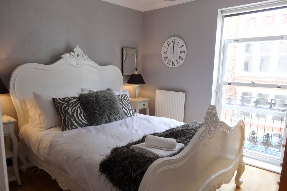 A bedroom with a white ornate bed, a round wall clock, a window with a view of buildings, and a radiator.