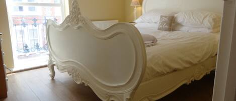 A bedroom with a white ornate bed, a nightstand, a lamp, and a window with a view of buildings.