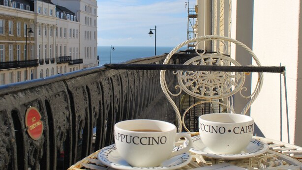 A balcony with a table set for two, featuring a newspaper and two cups labeled "PUCCINO" and "CAPPI." - Brighton Break - Burlington Beach Pad (Brighton)