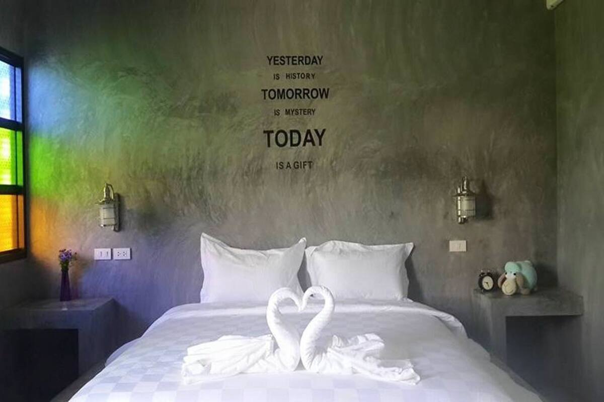 A bedroom with a bed, pillows, a wall with a motivational quote, a bedside table with a lamp, and a nightstand with a clock and a teddy bear.