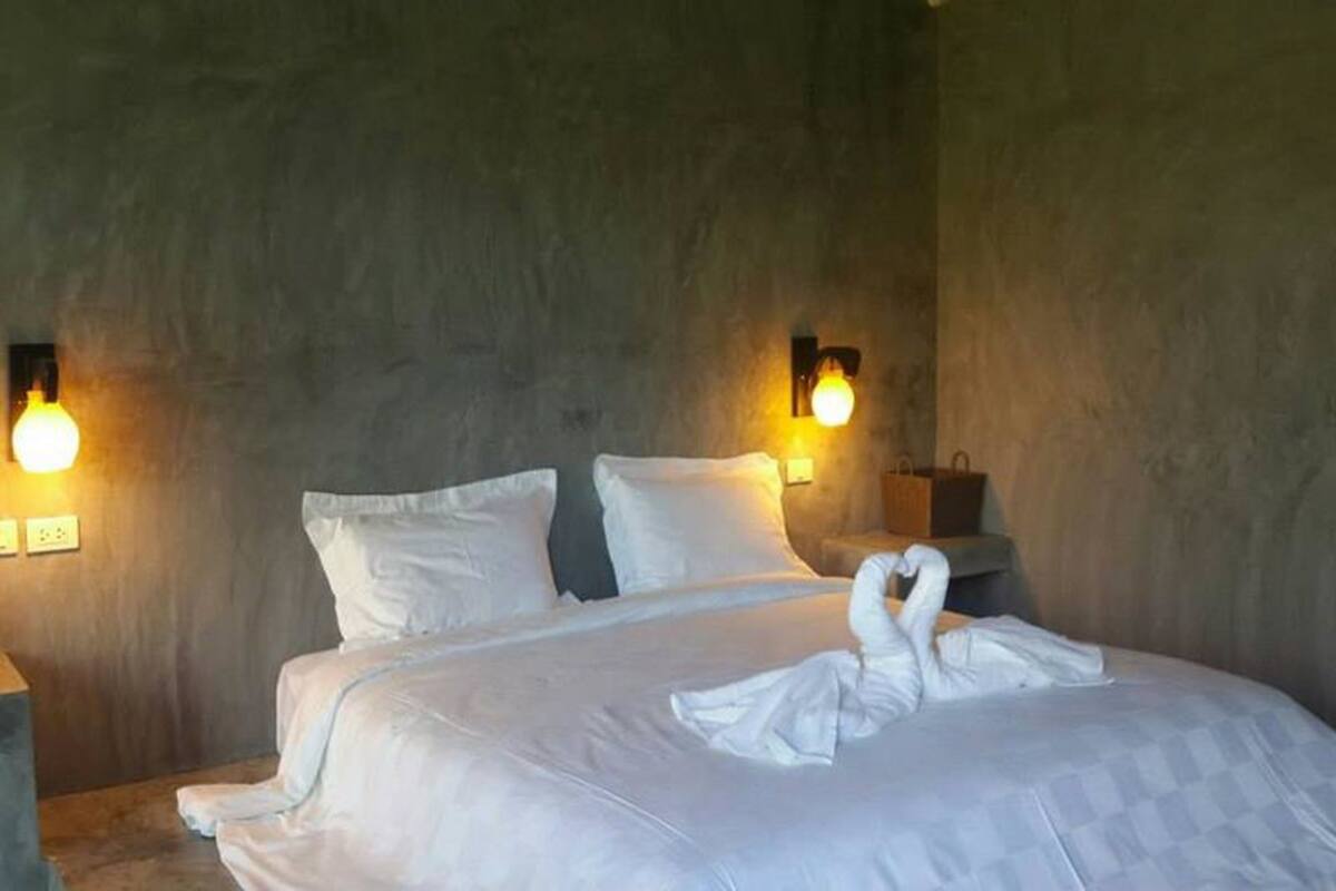 A neatly made bed with a swan-shaped pillow arrangement, two wall-mounted lamps, and a wooden nightstand with a bag.