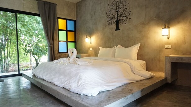 A modern bedroom with a large bed, a concrete platform, a tree mural, and a view of greenery through large windows. - Baanwaan Resort Hat Yai (Hat Yai)
