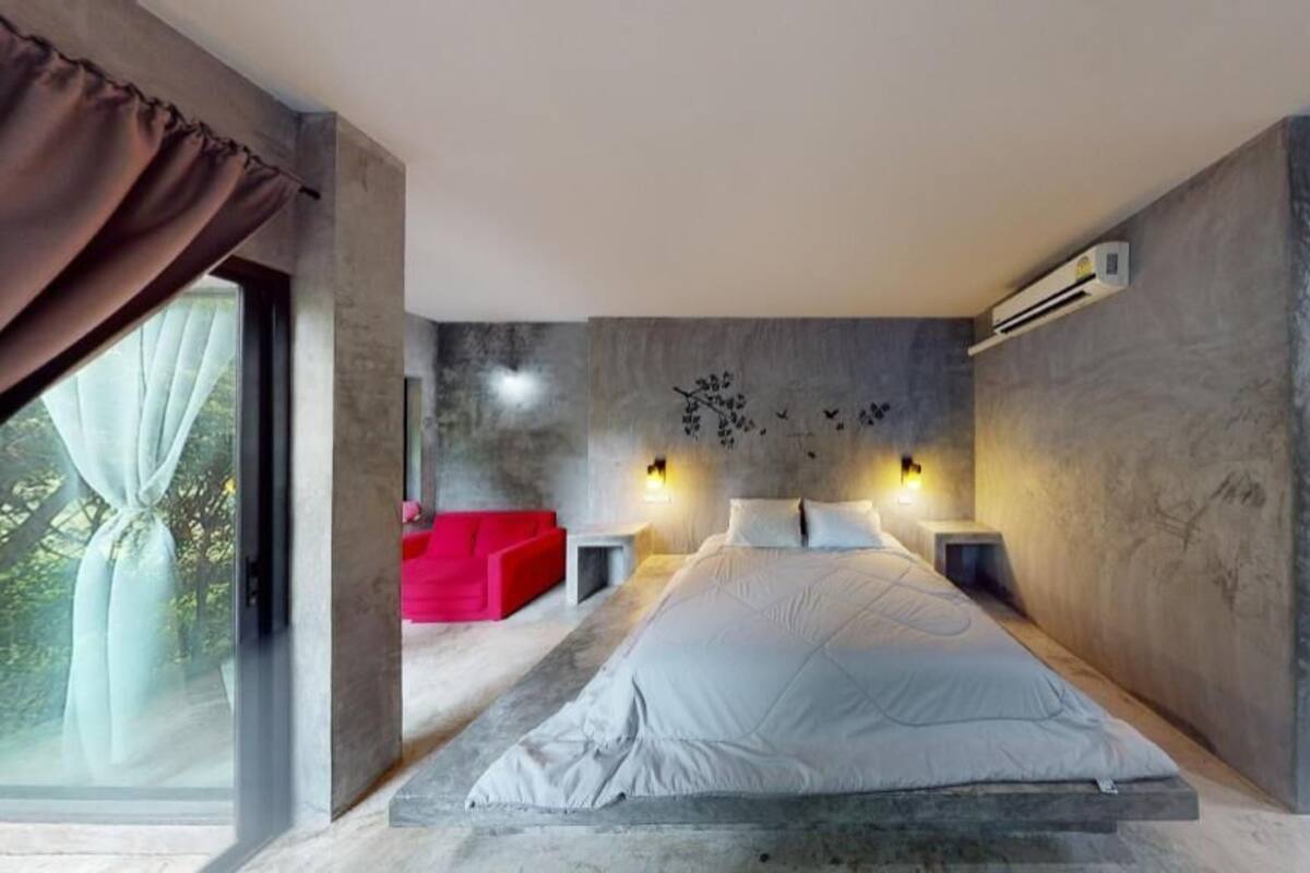 A modern bedroom with a large bed, a red sofa, and a view through a large window.