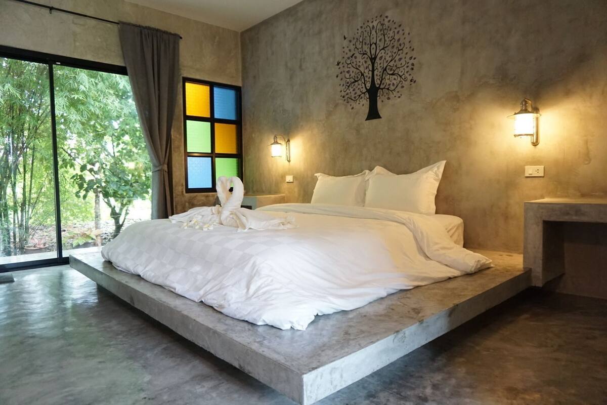 A modern bedroom with a large bed, a tree mural, and a view of greenery through the window.